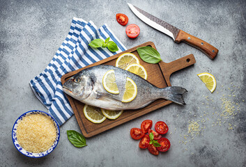 Fototapeta na wymiar Raw fish dorado with ingredients lemon, fresh basil, cut cherry tomatoes, uncooked rice on wooden cutting board with knife on rustic stone background top view, cooking healthy fish dorado concept