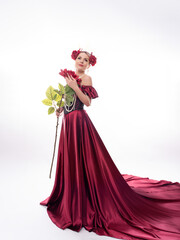 A model in a red satin dress in a crown with flowers and a pearl necklace, with a large red rose. A chic look. Full length photo on a white background