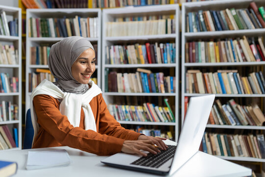 Young beautiful student in hijab studying online remotely in university library, woman typing on laptop keyboard searching for information and preparing for exam.