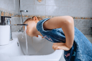 A little girl in a denim dress with a print leaned on a white ceramic sink to drink water from the tap in the bathroom.