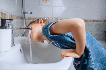 Foto op Plexiglas A little girl in a denim dress with a print leaned on a white ceramic sink to drink water from the tap in the bathroom. © Semachkovsky 