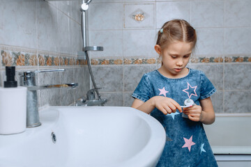 A little girl in a denim dress is standing in the bathroom near the ceramic sink and typing toothpaste on a brush.