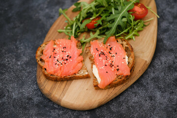 A wooden plate with salmon sandwiches, arugula, tomatoes on black background. Healthy food. Horizontal frame.