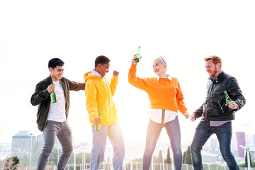 Group of multiracial friends dancing excitedly outdoors on spring day. Cheerful young people drinking beer and having fun. Colleagues celebrating party together. Concept of happiness and friendship. 
