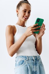 Millennial woman with beautiful and tanned skin looking at the phone screen in her hands and smiling in a white tank top and blue jeans against a white wall