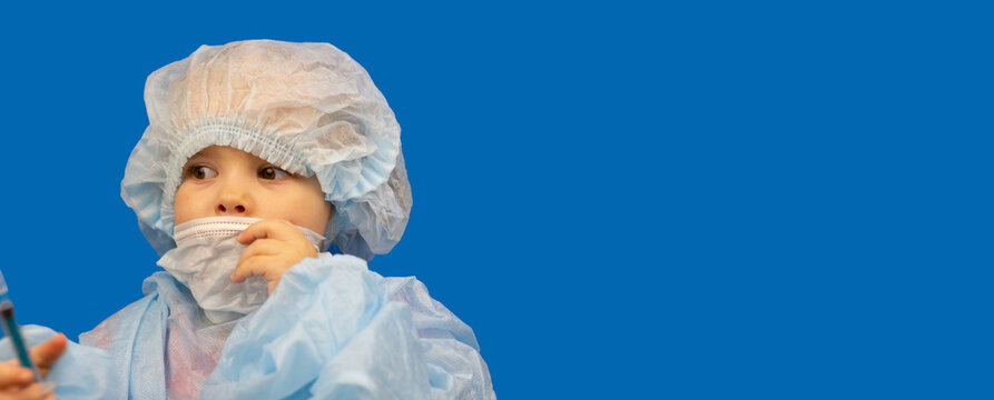 A small child in doctor's clothes on a blue background. Banner, place to insert. A cute baby in a medical mask, gown and cap on a blue background with empty space to insert.
