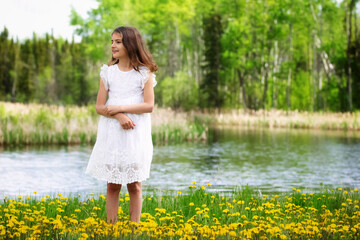 Girl in white dress is standing in the meadow with blooming dandelions in spring.