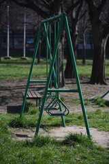 Empty old green metallic swing in the park with benches on a sunny day