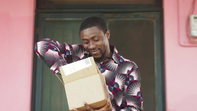 Happy African Man opening cardboard parcel box package with gifts purchased online, he received through postal shipping and delivery service. 