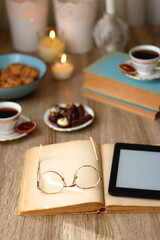 Books, reading glasses, e-reader, plate of chocolate pralines, bowl of cookies, cups of tea and lit candles on the table. Selective focus.