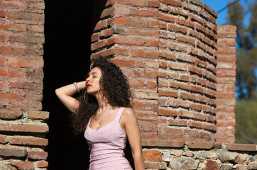 Beautiful brunette woman with curly hair among the ruins of an old abandoned building. The woman is dressed in an elegant pink and narrow dress.