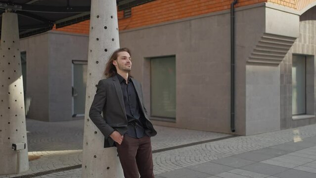 Handsome guy with beard and long hair is standing outdoor on office building background. He wears gray jacket, jeans. He looks to the side. man looking dreamy
