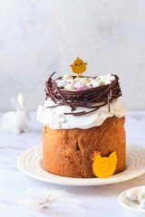 Easter cake covered with icing with a decor of a chocolate nest with eggs and a chicken topper on a white plate on a marble background. Easter table, treats, decor, traditions concept.