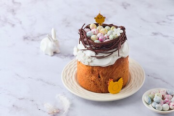 Easter cake covered with icing with a decor of a chocolate nest with eggs and a chicken topper on a white plate on a marble background. Easter table, treats, decor, traditions concept.