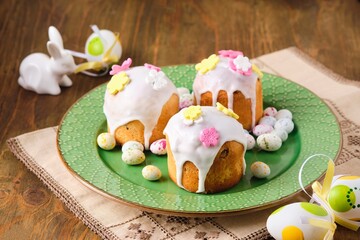 Mini Easter cakes covered with icing and decorated with sugar flowers on a green plate on a wooden background. Easter table, treats, decor, traditions concept.