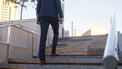 Unrecognizable young businessman climbs up backlit stone stairs in city. man in formal suit dark blue color with tablet in his hands walks forward, close up view of his feet in brown leather boots.