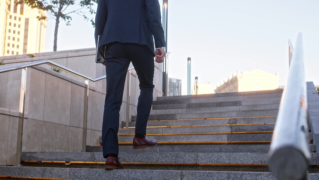 Unrecognizable young businessman climbs up backlit stone stairs in city. man in formal suit dark blue color with tablet in his hands walks forward, close up view of his feet in brown leather boots. - Powered by Adobe