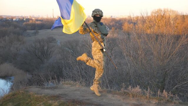 Ukrainian army man dancing funny with blue-yellow flag on hill at countryside. Happy male soldier in camouflage uniform celebrating victory of war between Ukraine and Russia. End of warfare in Europe