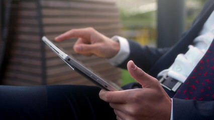 Close up successful businessman in suit sits outside in park and use tablet, writes with finger, transfers files. Unrecognizable man works on mobile computer outdoors. Lunch break between chores