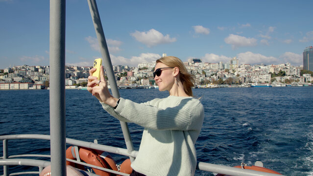 young woman in turquoise sweater takes selfie on smartphone while sailing on pleasure boat in middle of sea or strait. girl is photographed on phone at stern of ship.