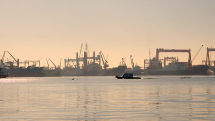 small white coast guard motorboat sails through bay with large port in background with cargo tankers. Sunset or dawn over port city in summer.
