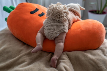 cute newborn baby in beige knitted rabbit costume is lying on orange plush pillow, infant is crawling on carrot toy, learning how to walk