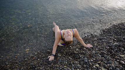 beautiful slender young woman in purple swimsuit lies on rocks on beach and waves wash over her body. girl is resting on stone beach look camera, half lying in water. Morning relaxation in pond.