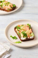 Smorrebrod, an open sandwich on rye bread with boiled salmon, fresh cucumber, cream cheese, apple and herbs on a ceramic plate on a light concrete background. Danish cuisine.