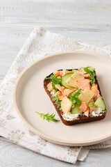 Smorrebrod, an open sandwich on rye bread with boiled salmon, fresh cucumber, cream cheese, apple and herbs on a ceramic plate on a light concrete background. Danish cuisine.