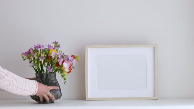 Female hands puts flowers in a vase. Wooden frame mockup in white room interior, empty mock-up with copy space. Light frame with a vase in the interior for design presentation.