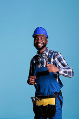Excited african american man in hard hat showing thumbs up as sign of approval. Optimistic builder happy at work wearing belt with tools and uniform, construction and renovations.