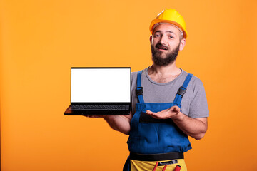 Cheerful builder showing laptop with white blank screen, holding pc with isolated copyspace display for advertisement in studio. Contractor worker using mockup template on computer.