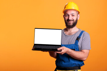 Smiling construction worker holding laptop with empty white screen for advertising renovation...