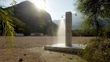 shower column stands alone on beach, water flows from shower. Mountains and sun in background....
