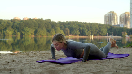 slender athletic young woman in tight gray tracksuit is doing workout and doing push-ups from her knees. beautiful girl in morning at dawn on river bank does sports outside in nature.