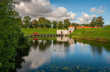 Copenhagen, Denmark - September 13, 2010: Green landscape of moat and ramparts with bridge and gate of south entrance to Kastellet, citadel, under blue cloudscape. Red roofs peep over