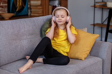 Happy child listens to music or audiobook at home on couch. Little school kid wearing headphones