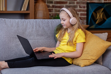 Little girl with headphones and laptop on sofa in room. Child is playing a video game.