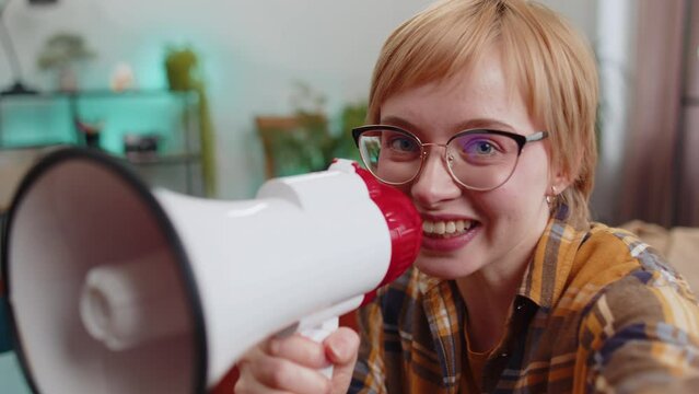 POV selphi portrait of young woman talking with megaphone, proclaiming news, loudly announcing advertisement, warning using loudspeaker to shout speech sitting on sofa. Girl at home room apartment