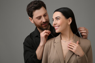 Man putting elegant necklace on beautiful woman against dark grey background