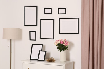 Empty frames hanging on white wall and chest of drawers with flowers indoors