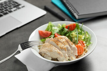 Bowl of tasty food, laptop, fork and notebooks on grey table. Business lunch