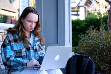teenage girl with a laptop sitting on the porch of house. She watching the news doing homework...