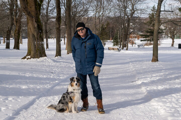 Fototapeta na wymiar St. John's, Newfoundland, Canada-April 2023: An older man dressed in winter clothes stands next to an Australian shepherd dog in a park. The ground is covered in white snow and there are large trees.