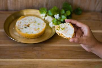 hand holding a piece of Ghewar or ghevar, which is a traditional sweet of rajasthan. It is made of milk, flour, ghee and sugar. Dry fruits and malai cream on top. It is served in brass plate.