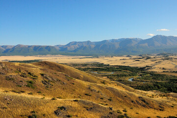 A view from the top of the hill to a valley yellowed by autumn at the foot of a mountain range with snow-capped peaks and a flowing meandering river below.