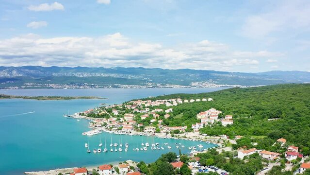 The aerial view of Malinska in Krk, Croatia displays a charming coastal town with a marina full of boats, a pristine beach lined with palm trees, and picturesque Mediterranean architecture surrounded 