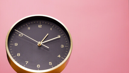 Front view of black table clock, gold dial on pink background, hour hand pointing one, minute hand pointing two.