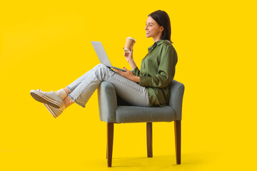 Beautiful woman with cup of coffee using laptop in soft armchair on yellow background