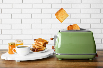 Crispy bread slices in modern toaster, cup of coffee and toasts with jam on wooden table near white...